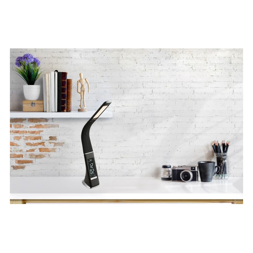 Stolná lampa svieti na bielom stole pred tehlovou stenou. Lampa má zakrivený tvar, digitálny displej zobrazujúci čas 12:02 a je umiestnená na stole s fotoaparátom, pohárom a písacími potrebami. Na poličke za lampou sú knihy a dekorácia.