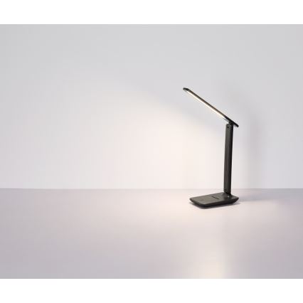 Stolná lampa svieti na svetlošedom stole. Lampa je skladacia, tmavosivá, s tenkým ramenom. Svetlo smeruje doprava a mierne nahor. Pozadie tvorí svetlá stena.