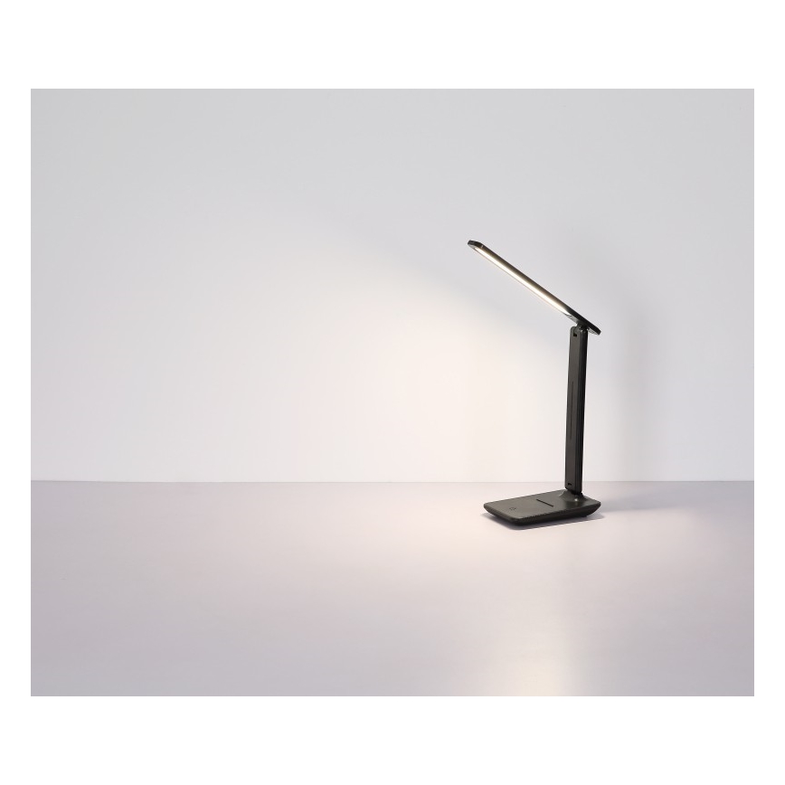 Stolná lampa svieti na svetlošedom stole. Lampa je skladacia, tmavosivá, s tenkým ramenom. Svetlo smeruje doprava a mierne nahor. Pozadie tvorí svetlá stena.