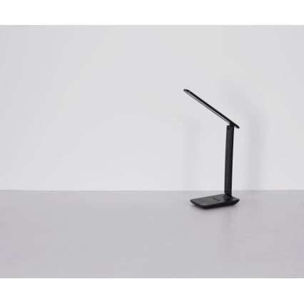 Čierna stolová lampa s ohybným ramenom stojí na svetlom stole pred svetlou stenou. Lampa je zložená a smeruje doprava.