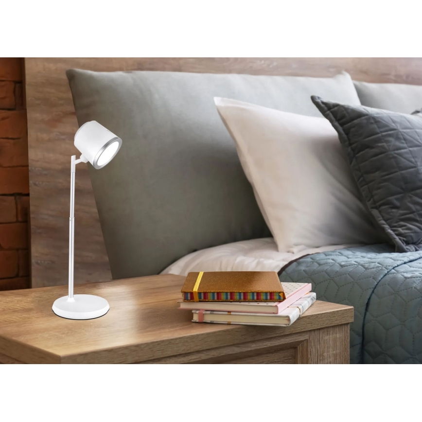 Biela stolová lampa s nastaviteľným ramenom stojí na nočnom stolíku. Lampa osvetľuje tri knihy. Stolík sa nachádza v spálni vedľa postele.