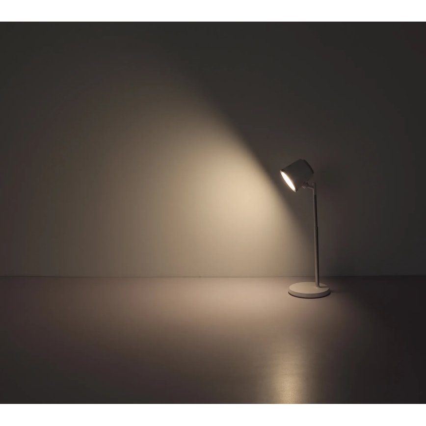 Stolová lampa svieti. Je umiestnená na tmavej podlahe pred svetlou stenou. Lampa je biela a kovová. Svetlo z lampy smeruje doprava a osvetľuje stenu.