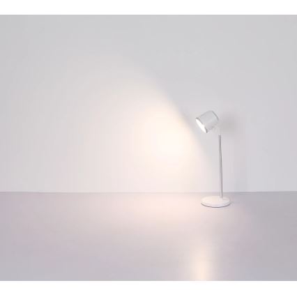 Biela stolová lampa s nastaviteľným ramenom svieti na bielu stenu. Lampa stojí na svetlej podlahe.