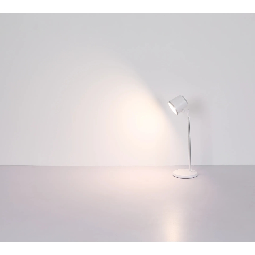 Biela stolová lampa s nastaviteľným ramenom svieti na bielu stenu. Lampa stojí na svetlej podlahe.