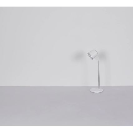 Biela stolná lampa s nastaviteľným ramenom stojí na svetlosivej ploche pred bielou stenou. Lampa je v zapnutom stave a smeruje doprava.