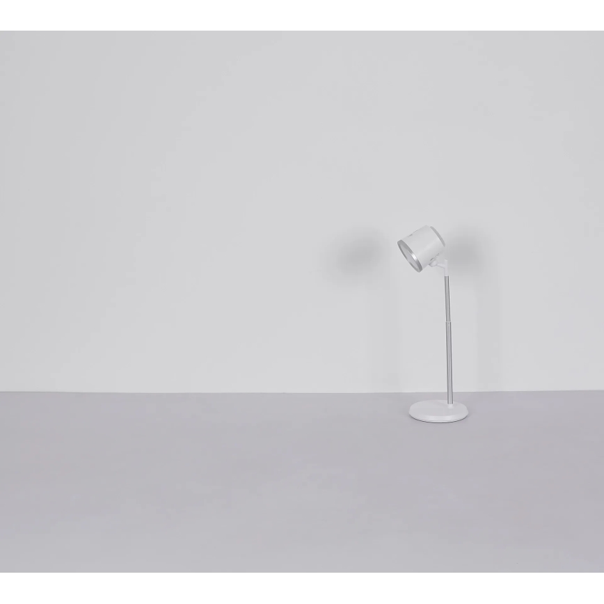 Biela stolná lampa s nastaviteľným ramenom stojí na svetlosivej ploche pred bielou stenou. Lampa je v zapnutom stave a smeruje doprava.