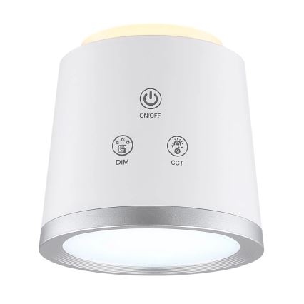 Biela valcovitá lampa svieti. Na tele sú tlačidlá: ON/OFF, DIM a CCT. Lampa má strieborný okraj.