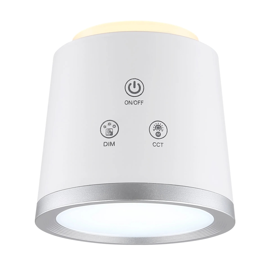 Biela valcovitá lampa svieti. Na tele sú tlačidlá: ON/OFF, DIM a CCT. Lampa má strieborný okraj.
