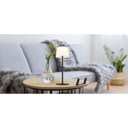 Stolová lampa s bielym tienidlom a čiernou kovovou nohou svieti na drevenom stolíku. Stolík stojí pred sivou sedačkou s vlnenou dekou. V blízkosti lampy je váza s rastlinou.