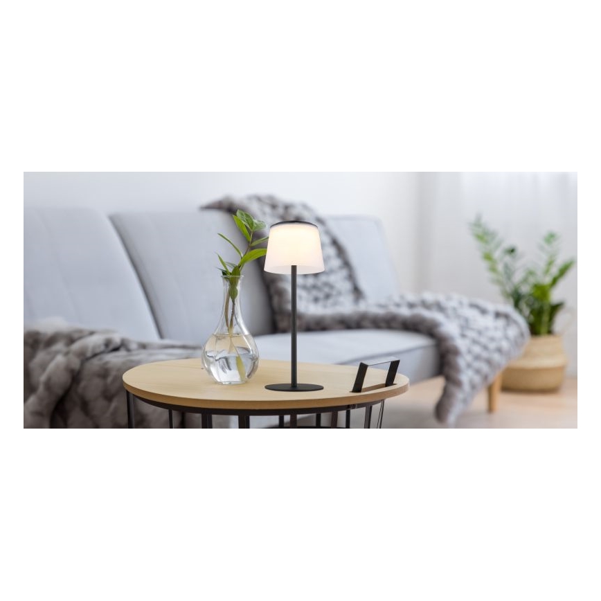 Stolová lampa s bielym tienidlom a čiernou kovovou nohou svieti na drevenom stolíku. Stolík stojí pred sivou sedačkou s vlnenou dekou. V blízkosti lampy je váza s rastlinou.