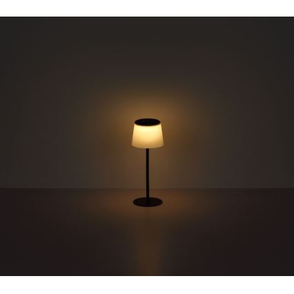 Stolová lampa s valcovým tienidlom svieti v tmavej miestnosti na tmavej podlahe. Lampa je na tenkej, tmavej nohe s kruhovou základňou.
