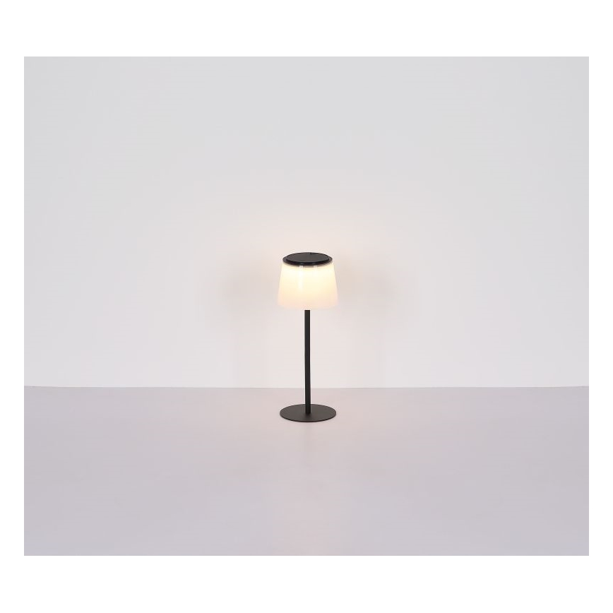 Stolová lampa s valcovým tienidlom zo svetlého materiálu a čiernym kovový podstavcom svieti na svetlom povrchu.