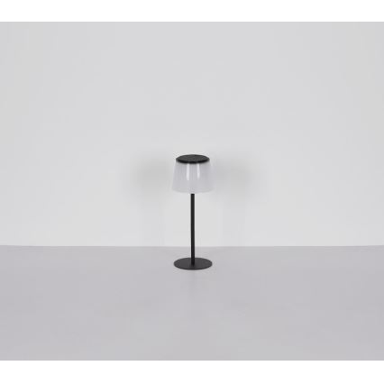 Stolová lampa s mliečnym tienidlom a čiernym kovovým stojanom stojí na svetlej ploche pred bielou stenou.