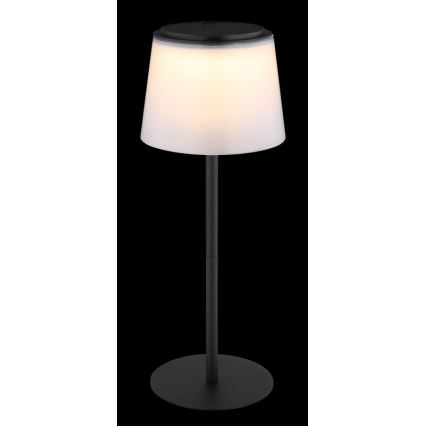 Solárna stolová lampa svieti. Má biely tienidlo a čierny kovový stojan s okrúhlou základňou. Lampa je umiestnená na tmavom pozadí.