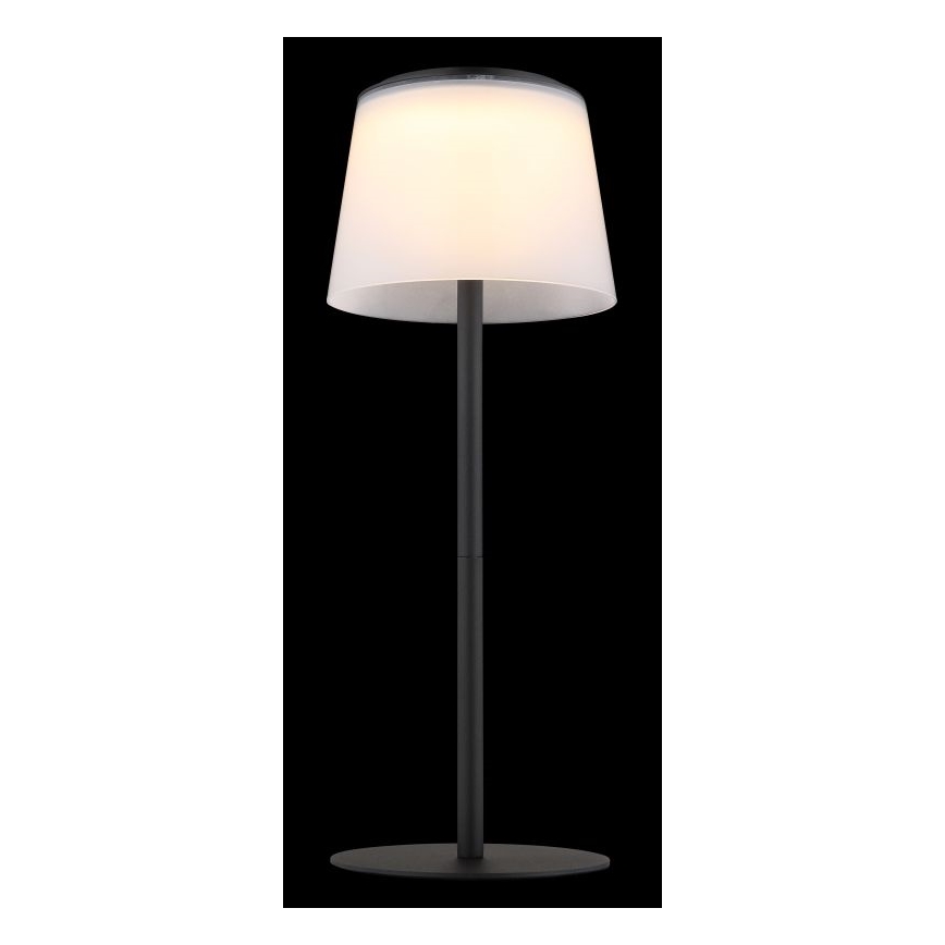 Stolová lampa svieti. Má biely tienidlo a čierny kovový stojan s okrúhlou základňou. Je umiestnená na tmavom pozadí.