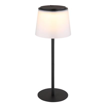 Stojacia lampa svieti. Má čierny kovový stojan a biely tienidlo. Na vrchu tienidla je tmavý kryt, pravdepodobne slnečný panel alebo ovládací prvok.