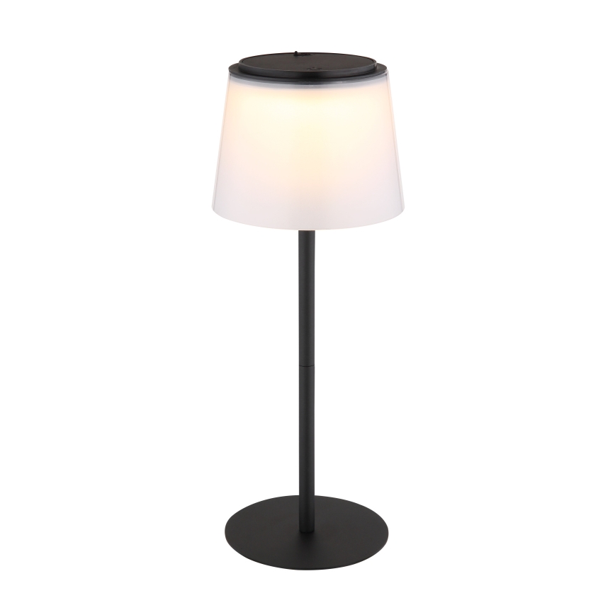 Stojacia lampa svieti. Má čierny kovový stojan a biely tienidlo. Na vrchu tienidla je tmavý kryt, pravdepodobne slnečný panel alebo ovládací prvok.