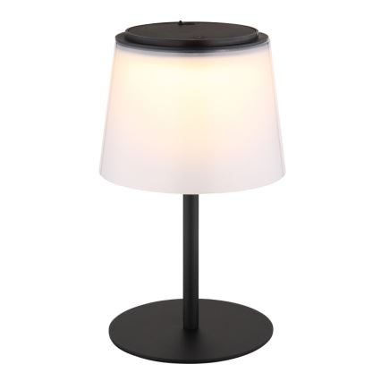 Stolová lampa s bielym tienidlom a čiernou kovovou nohou svieti. Vrchná časť tienidla má integrovaný solárny panel. Lampa stojí na kruhovej čiernej základni.