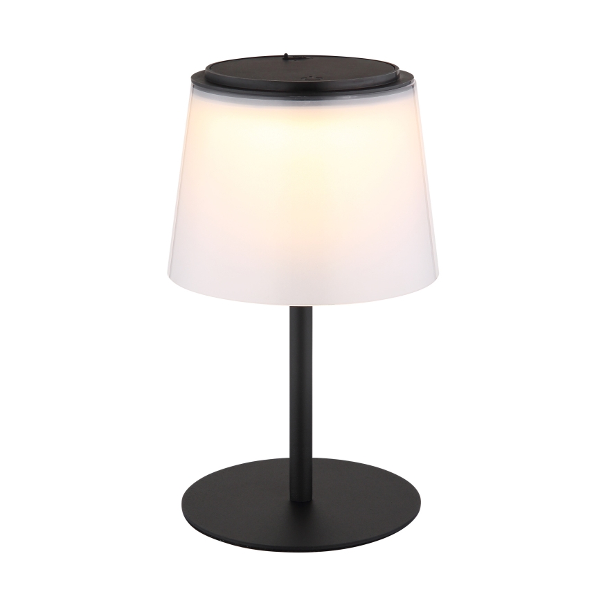 Stolová lampa s bielym tienidlom a čiernou kovovou nohou svieti. Vrchná časť tienidla má integrovaný solárny panel. Lampa stojí na kruhovej čiernej základni.