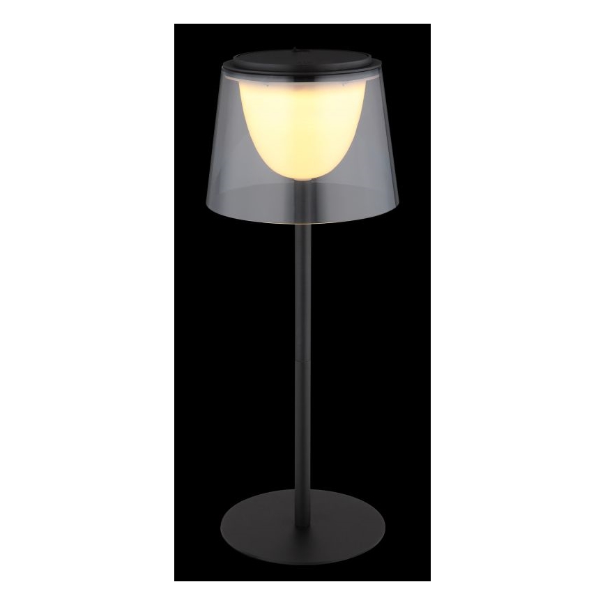 Stolová lampa svieti. Má čierny kovový stojan a priesvitný tienidlo s teplým svetlom. Lampa stojí na tmavom pozadí.