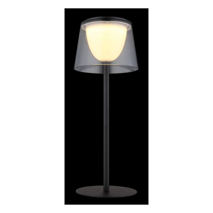 Solárna lampa svieti. Má čierny kovový stojan a priesvitný tienidlo zo skla, ktoré obklopuje teplé biele svetlo. Je umiestnená na čiernom podklade.
