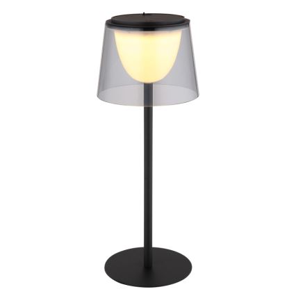 Stojacia lampa svieti. Má čierny kovový stojan a priesvitný tienidlo z plastu nad teplým svetlom. Lampa stojí na kruhovom podstavci.