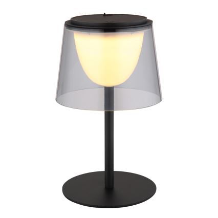 Stolová lampa svieti. Má čierny kovový stojan a tienidlo zo skleneného zvonu, vnútri svieti teplá biela LED dióda. Celok stojí na čiernej kruhovej podstave.