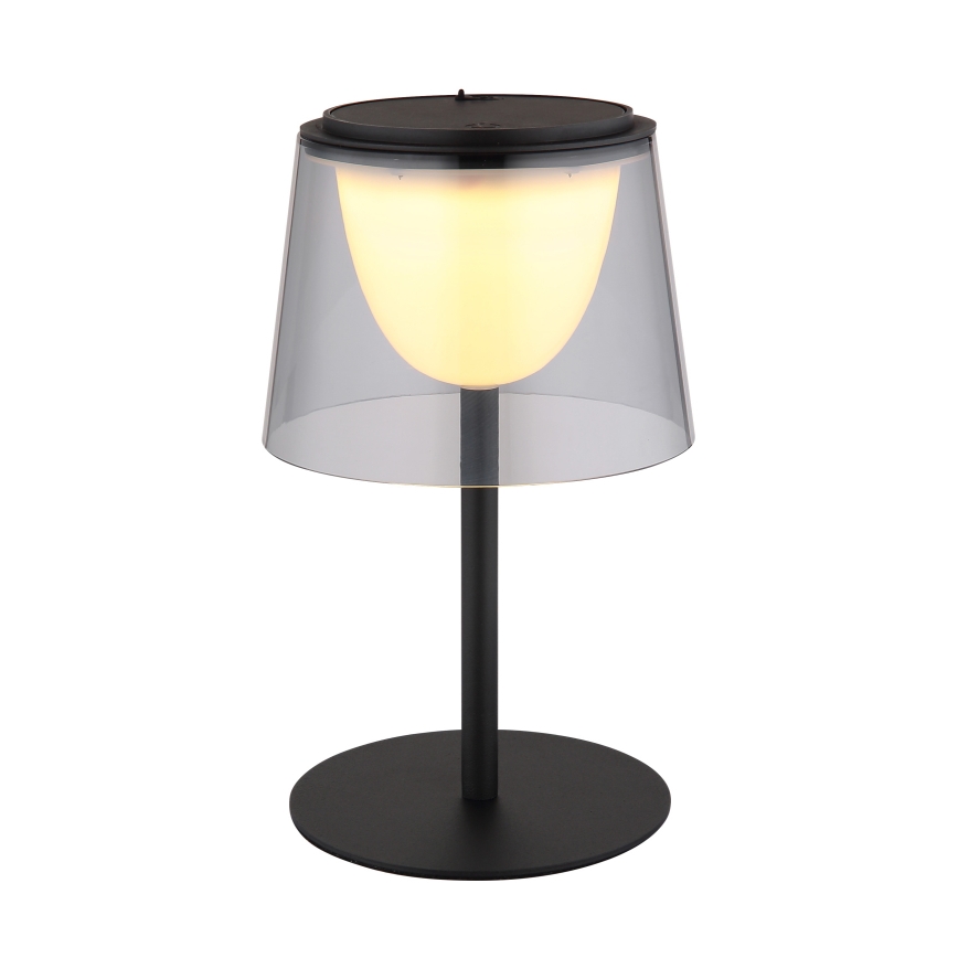 Stolová lampa svieti. Má čierny kovový stojan a tienidlo zo skleneného zvonu, vnútri svieti teplá biela LED dióda. Celok stojí na čiernej kruhovej podstave.
