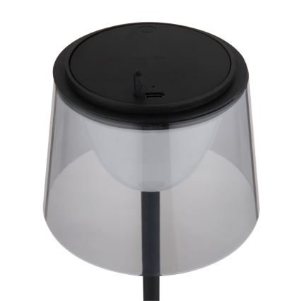 Stolová lampa s priesvitným tienidlom a čiernym podstavcom svieti. Na vrchu je čierny kryt s USB portom. Lampa je umiestnená na bielom pozadí.