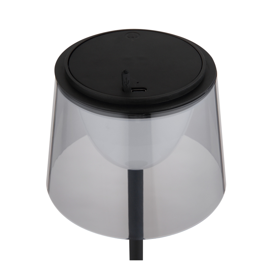 Stolová lampa s priesvitným tienidlom a čiernym podstavcom svieti. Na vrchu je čierny kryt s USB portom. Lampa je umiestnená na bielom pozadí.