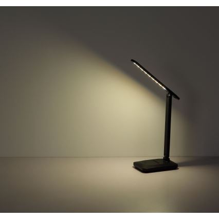 Čierna stolová lampa s LED osvetlením svieti na svetlohnedú stenu a plochu. Lampa je zložená a umiestnená na svetlej ploche.