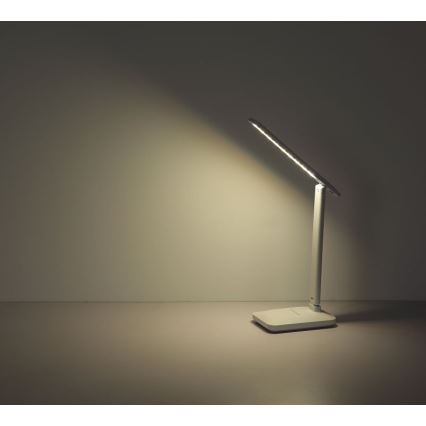 Stolná lampa s LED osvetlením svieti na stôl. Lampa je biela, má štíhly dizajn s dlhým, tenkým ramenom. Nachádza sa na svetlom povrchu pred svetlou stenou.