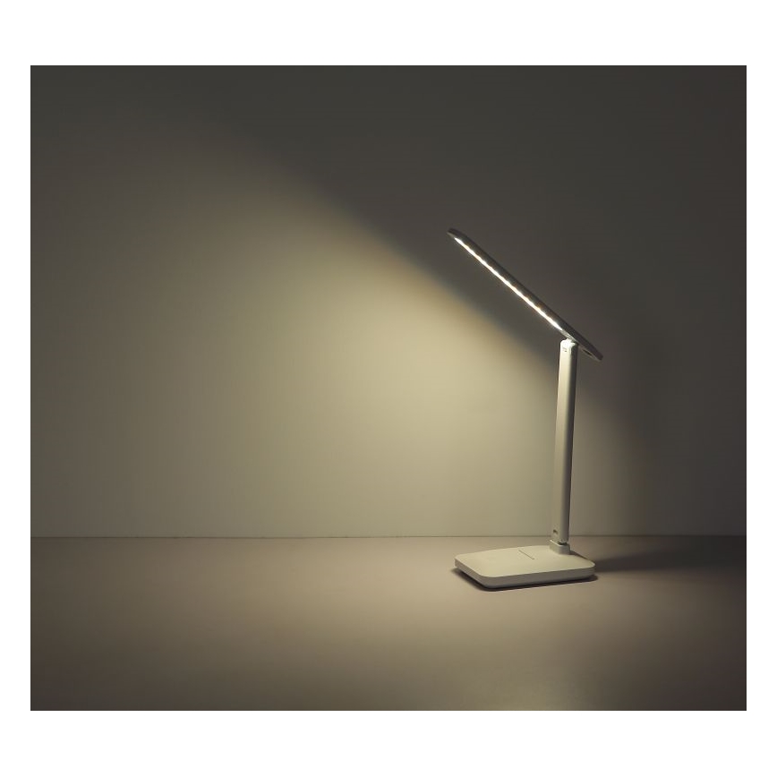 Stolná lampa s LED osvetlením svieti na stôl. Lampa je biela, má štíhly dizajn s dlhým, tenkým ramenom. Nachádza sa na svetlom povrchu pred svetlou stenou.
