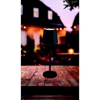Čierna stolová lampa s kužeľovitým tienidlom stojí na drevenom stole. Lampa svieti. Na pozadí je rozostrený záhradný dom s vonkajším osvetlením.