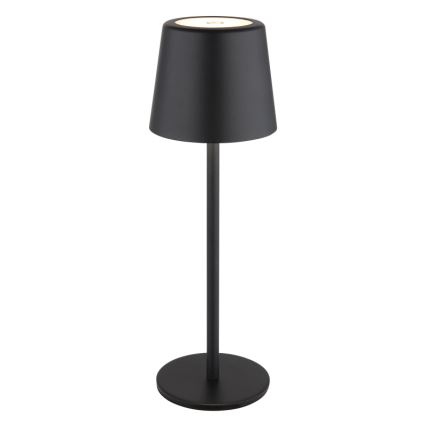 Čierna stolová lampa s valcovým tienidlom svieti. Stredne vysoká, s úzkym stĺpikom a kruhovou podstavou. Na vrchu tienidla je vidieť malý svetelný zdroj.