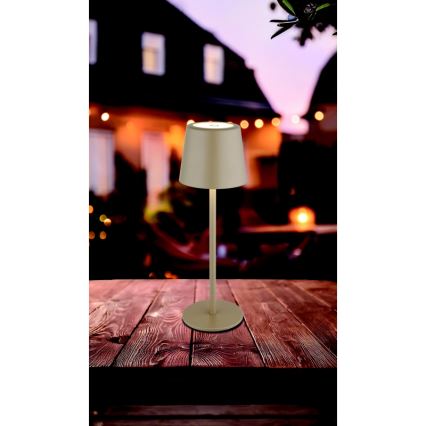 Stolová lampa s béžovým tienidlom a kovovou nohou stojí na tmavom drevenom stole. Svieti v pozadí vidieť rozostrené domy s vonkajším osvetlením.