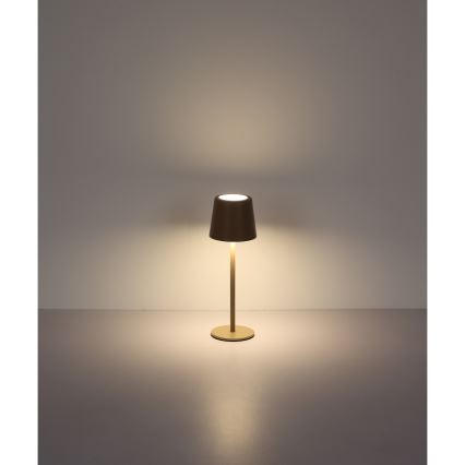 Stolová lampa svieti. Má valcovitý tienidlo a vysokú, úzku nohu na kruhovej základni. Stojí na svetlej ploche pred svetlou stenou.