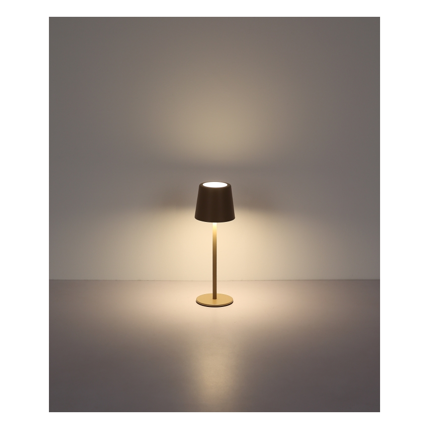 Stolová lampa svieti. Má valcovitý tienidlo a vysokú, úzku nohu na kruhovej základni. Stojí na svetlej ploche pred svetlou stenou.