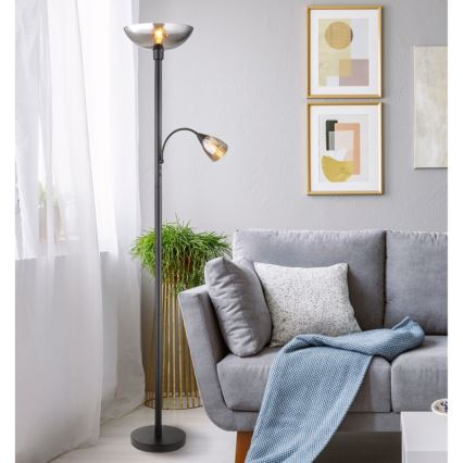 Dvojramenná stojacia lampa s tienidlami z dymovateného skla stojí v obývacej izbe vedľa sivej pohovky. Lampa je čierna, má jeden väčší a jeden menší svetelný zdroj.