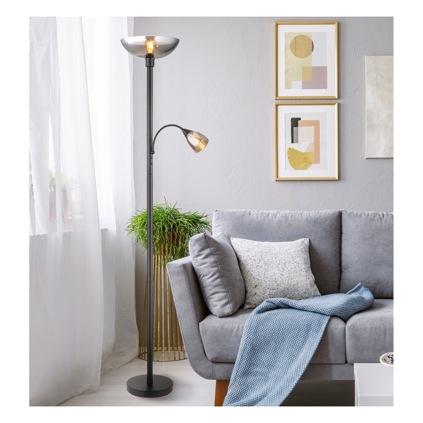 Dvojramenná stojacia lampa s tienidlami z dymovateného skla stojí v obývacej izbe vedľa sivej pohovky. Lampa je čierna, má jeden väčší a jeden menší svetelný zdroj.