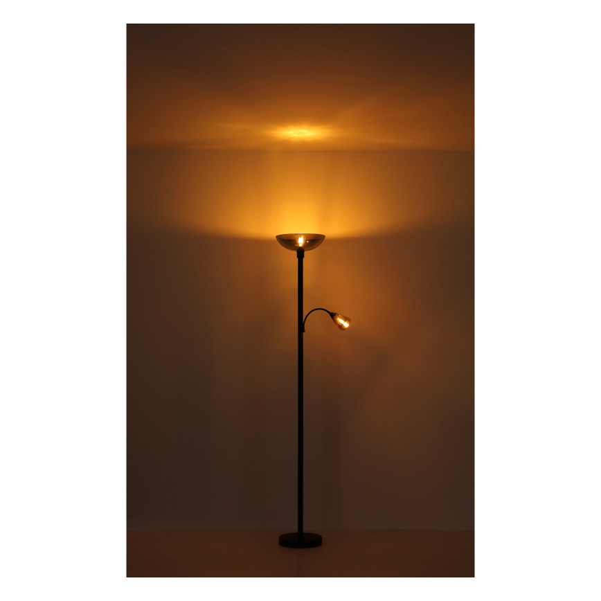 Stojacia lampa s dvoma svetlami svieti. Hlavné svetlo je široké, svieti nahor a dole. Vedľajšie svetlo je malé, na ohybnom rameni. Lampa stojí na podlahe pred svetlou stenou.