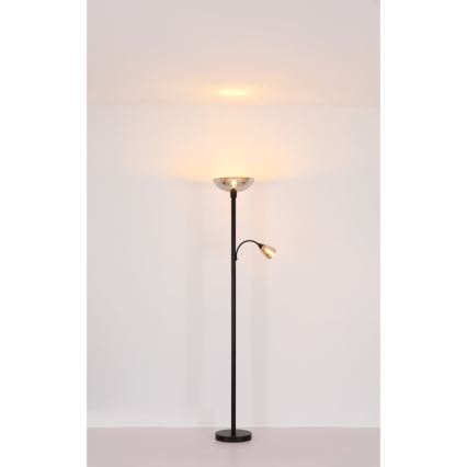 Stojacia lampa s dvoma svetlami svieti v svetlej miestnosti. Hlavné svetlo má široký, miskovitý tienidlo, druhé menšie svetlo je na výsuvnom rameni. Lampa má čiernu kovovu nohu a stojí na svetlej podlahe.