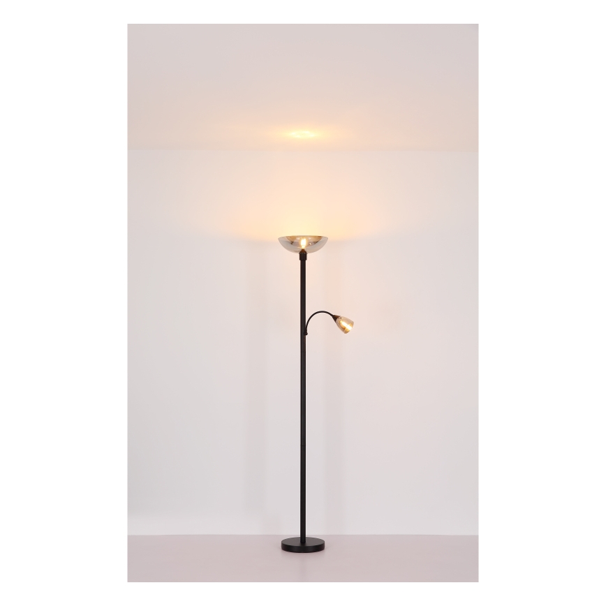 Stojacia lampa s dvoma svetlami svieti v svetlej miestnosti. Hlavné svetlo má široký, miskovitý tienidlo, druhé menšie svetlo je na výsuvnom rameni. Lampa má čiernu kovovu nohu a stojí na svetlej podlahe.