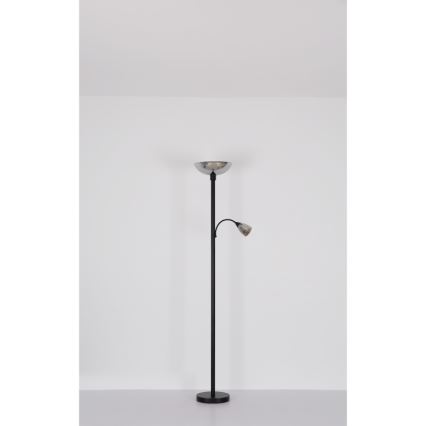 Stojacia lampa s dvoma svetlami stojí v bielej miestnosti. Hlavné svetlo má široký strieborný tienidlo, menšie svetlo je pripevnené na výsuvnom rameni. Lampa má čiernu kovové telo.