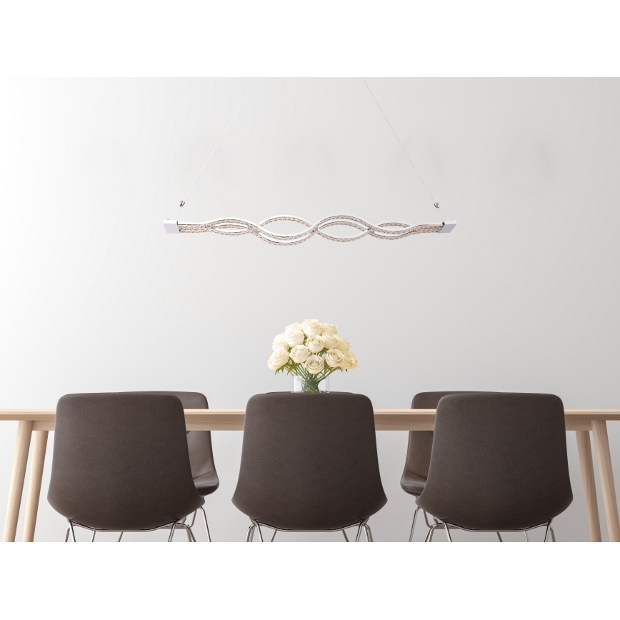 Svietidlo visí nad jedálenským stolom. Svietidlo má tvar vlniek z LED diód. Stôl je obklopený hnedými stoličkami. Na stole je váza s bielymi ružami.