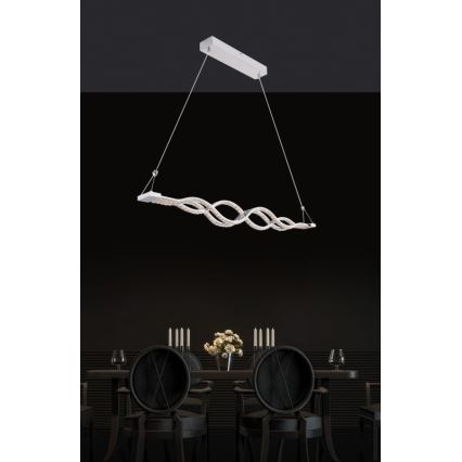 Svietidlo s vlnovitým tvarom visí nad jedálenským stolom. Svietidlo je zavesené na dvoch tenkých kábloch a je osvetlené LED diódami. Stojí nad tmavou jedálňou s čiernymi stoličkami.