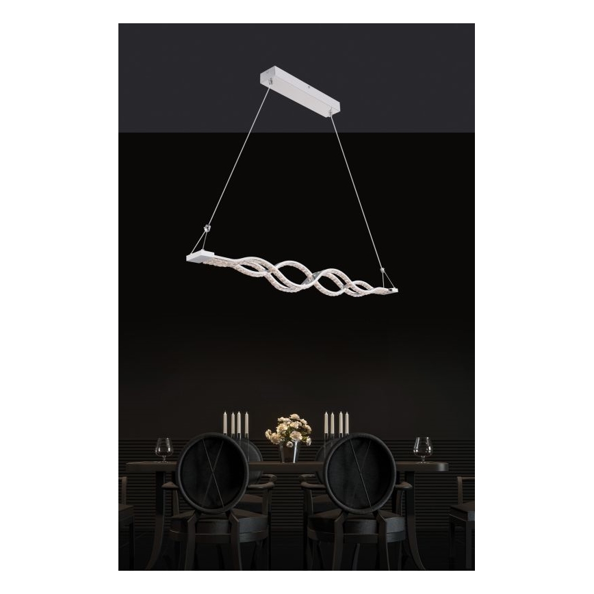 Svietidlo s vlnovitým tvarom visí nad jedálenským stolom. Svietidlo je zavesené na dvoch tenkých kábloch a je osvetlené LED diódami. Stojí nad tmavou jedálňou s čiernymi stoličkami.