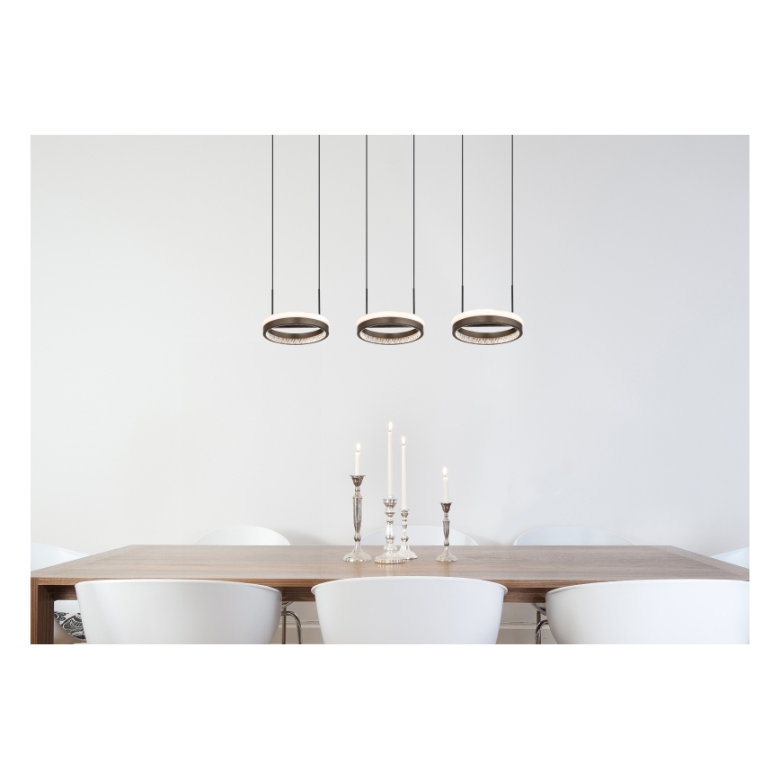 Trojica kruhových závesných svietidiel s LED osvetlením visí nad dreveným jedálenským stolom s bielymi stoličkami a striebornými svietnikmi.