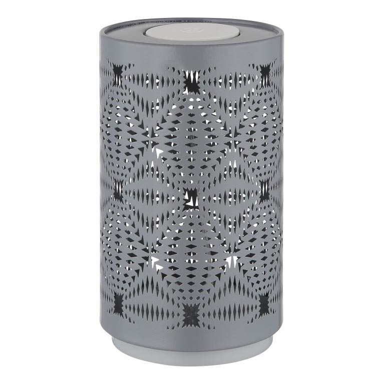Sivá kovová stolná lampa valcovitého tvaru s vyrezávaným geometrickým vzorom stojí na bielom podklade. Lampa je umiestnená na svetlom podstavci.