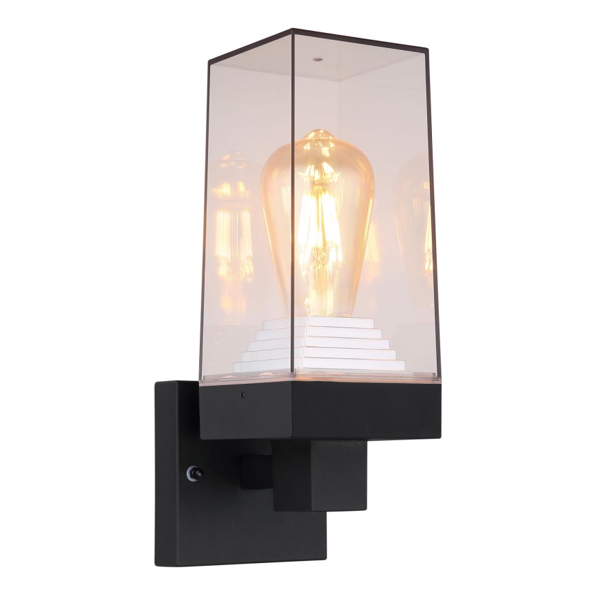 Nástená lampa svieti. Je čiernej farby so skleneným štvorcovým krytom, v ktorom je umiestnená žiarovka tvaru Edisonky. Lampa je pripevnená na stenu.